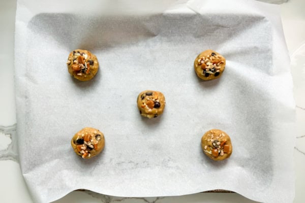 The ultimate sweet and salty kitchen sink cookies with a brown butter base, gooey caramel, double chocolate, pretzels, chips, and a flaky sea salt finish. Five balls of kitchen sink cookie dough topped with chocolate chips and sprinkles are arranged on a parchment-lined baking sheet, ready to be baked. The tray is placed on a white marble surface.