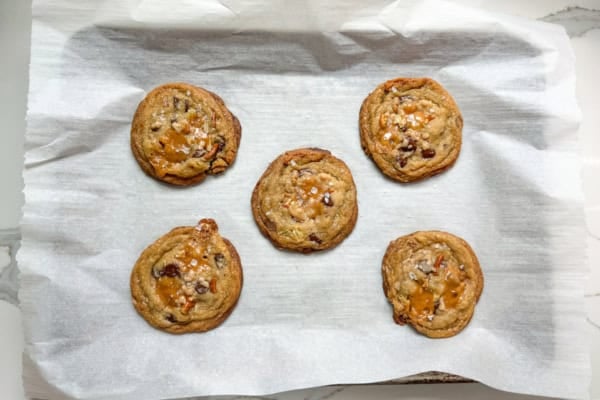 The ultimate sweet and salty kitchen sink cookies with a brown butter base, gooey caramel, double chocolate, pretzels, chips, and a flaky sea salt finish. Five brown butter kitchen sink cookies with pools of caramel and sprinkles of flaky sea salt on top are arranged on a parchment-lined baking tray, viewed from above on a light-colored countertop.