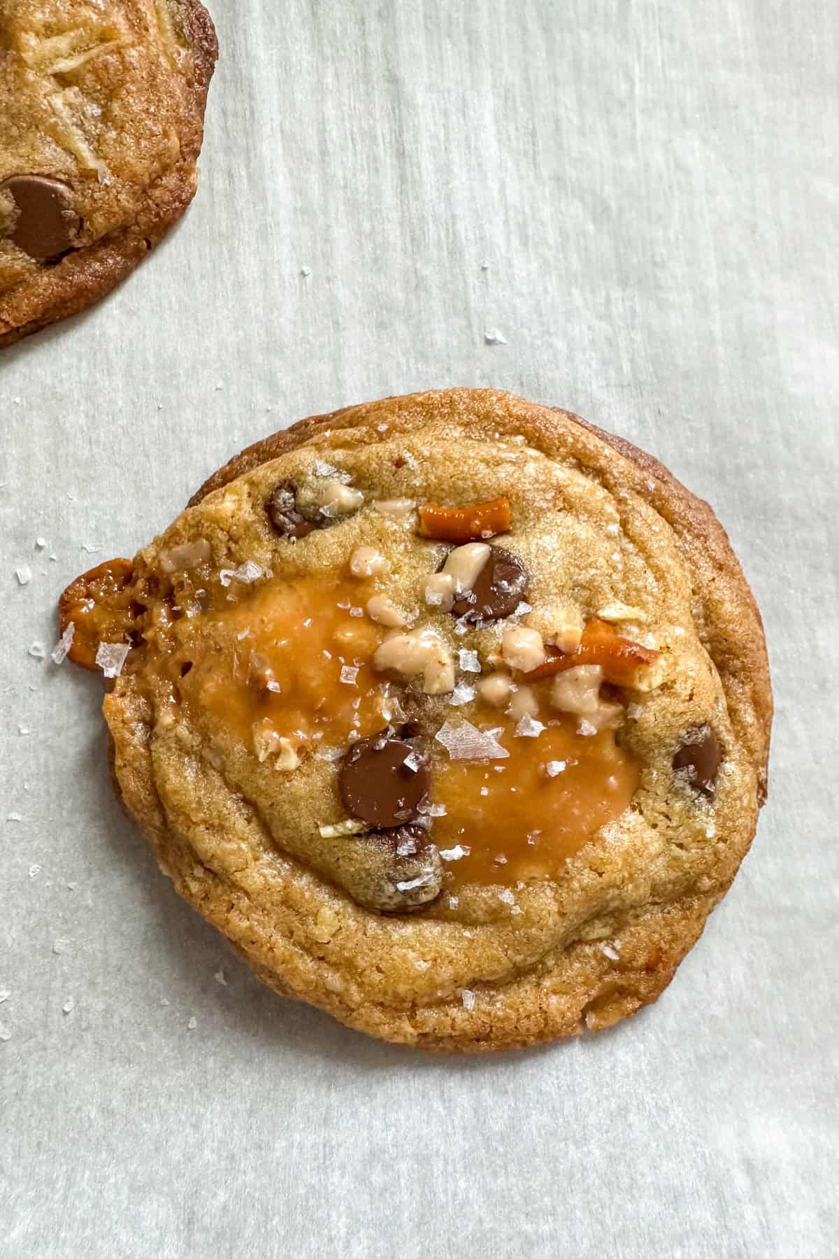 The ultimate sweet and salty kitchen sink cookies with a brown butter base, gooey caramel, double chocolate, pretzels, chips, and a flaky sea salt finish. A close-up of a panera kitchen sink cookie with a gooey caramel center, chocolate chips, pretzel pieces, and a sprinkle of flaky sea salt on top, resting on parchment paper.