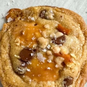 Close-up of a brown butter kitchen sink cookie with chocolate chips, caramel pieces, and flaky sea salt on top.