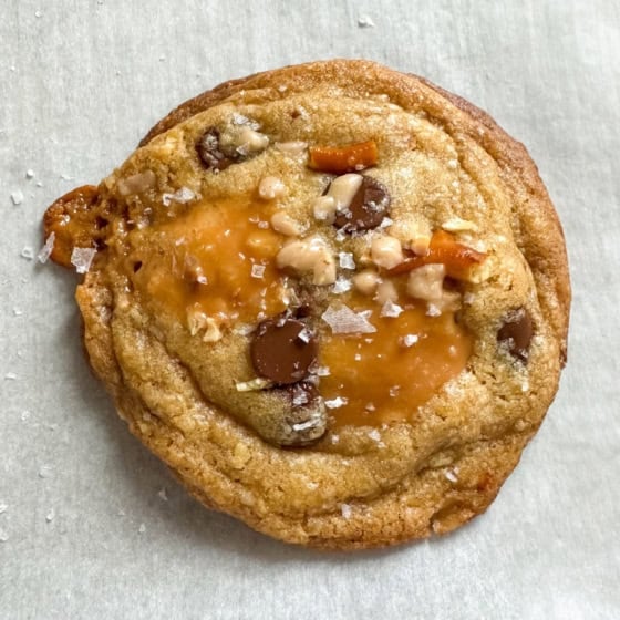 A close-up of a golden-brown Kitchen Sink Cookie with chocolate chips, caramel, pretzels, toffee bits, and flaky sea salt, resting on a sheet of parchment paper.