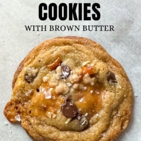 A close-up of a brown butter kitchen sink cookies with chocolate chips and caramel pieces topped with flaky sea salt on parchment paper.