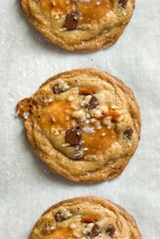 Three everything but the kitchen sink cookies with brown butter, chocolate chips, pretzels, and pools of caramel are topped with sea salt and arranged in a vertical row on parchment paper.