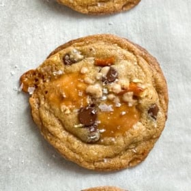 Three everything but the kitchen sink cookies with brown butter, chocolate chips, pretzels, and pools of caramel are topped with sea salt and arranged in a vertical row on parchment paper.