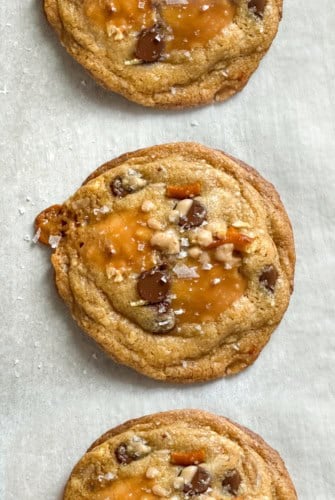 Three everything but the kitchen sink cookies with brown butter, chocolate chips, pretzels, and pools of caramel are topped with sea salt and arranged in a vertical row on parchment paper.