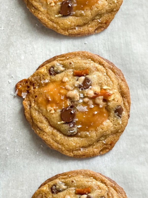 Three everything but the kitchen sink cookies with brown butter, chocolate chips, pretzels, and pools of caramel are topped with sea salt and arranged in a vertical row on parchment paper.