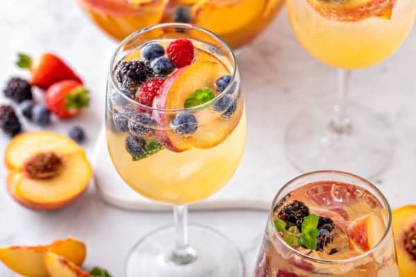 White Peach Sangria recipe is made with crisp white wine, peach nectar, schnapps and fresh fruit. The easiest summer cocktail you'll make all season! Glasses of white peach sangria garnished with peach slices, blueberries, blackberries, strawberries, and mint, with fresh fruit scattered around on a white surface.