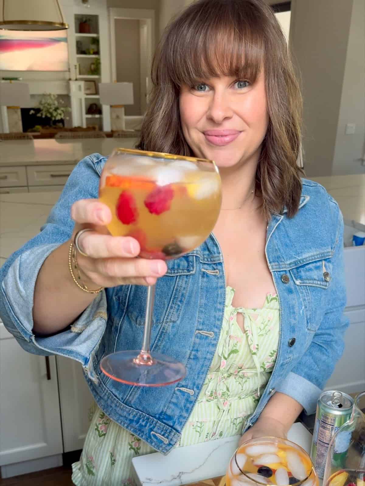 White Peach Sangria recipe is made with crisp white wine, peach nectar, schnapps and fresh fruit. The easiest summer cocktail you'll make all season! A woman with shoulder-length brown hair and bangs, wearing a denim jacket over a light green dress, smiles and holds up a large round glass of iced fruit punch in a bright kitchen.