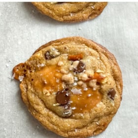 A close-up of a brown butter kitchen sink cookie with chocolate chips, pretzel pieces, caramel, and salt flakes on parchment paper.