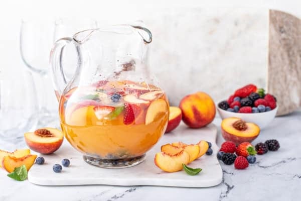 White Peach Sangria recipe is made with crisp white wine, peach nectar, schnapps and fresh fruit. The easiest summer cocktail you'll make all season! A glass pitcher filled with refreshing white peach sangria, garnished with slices of peaches, strawberries, and whole berries. Fresh fruit and a bowl of mixed berries are arranged beside it on a marble surface.