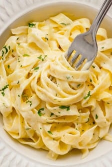 A close-up of creamy homemade Alfredo Sauce on fettuccine pasta garnished with chopped parsley in a white bowl, with a fork twirling some noodles.