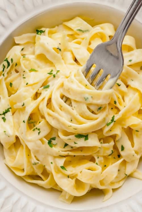 A close-up of creamy homemade Alfredo Sauce on fettuccine pasta garnished with chopped parsley in a white bowl, with a fork twirling some noodles.