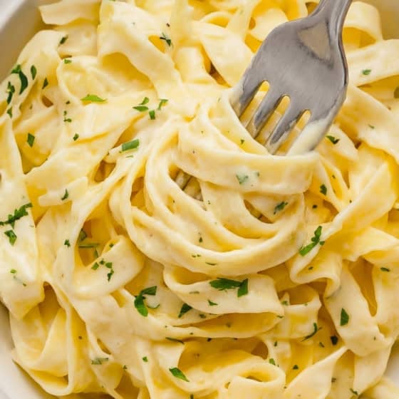 A close-up of creamy homemade Alfredo Sauce on fettuccine pasta garnished with chopped parsley in a white bowl, with a fork twirling some noodles.