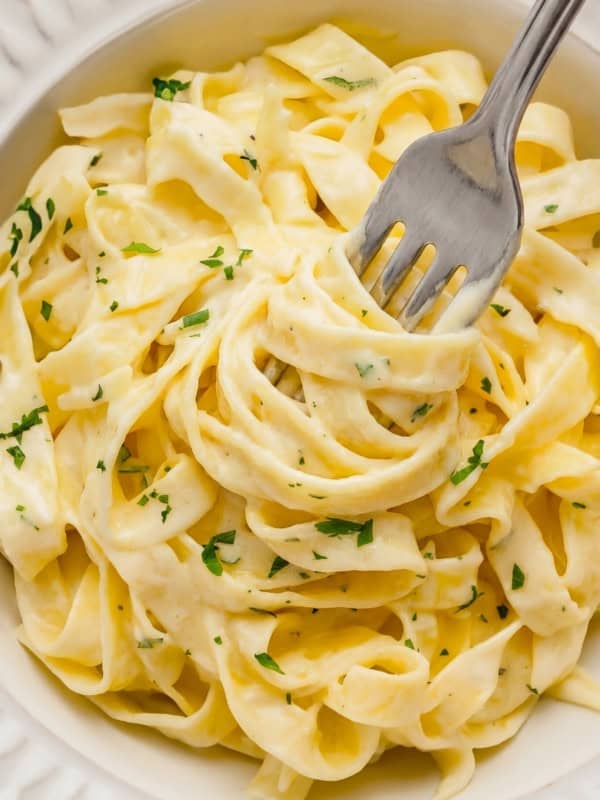 A close-up of creamy homemade Alfredo Sauce on fettuccine pasta garnished with chopped parsley in a white bowl, with a fork twirling some noodles.