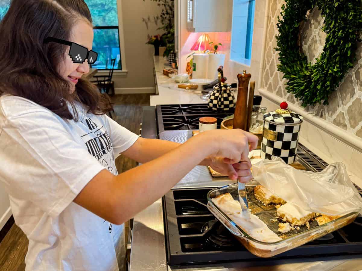 These lemon brownies are thick, chewy, packed with real lemon flavor, and topped with a sweet-tart glaze. Ready for the oven in 10 minutes! A girl wearing pixelated sunglasses cutting lemon brownies from a glass baking dish in a bright kitchen.