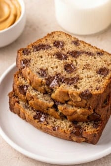 Three slices of chocolate chip peanut butter banana bread stacked on a white plate, with a bowl of peanut butter and a glass of milk in the background.