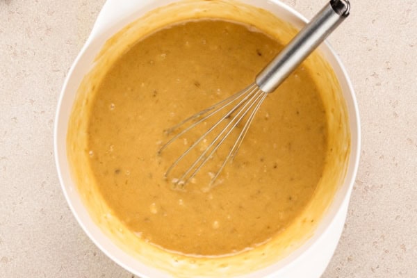 This Peanut Butter Banana Bread actually tastes like peanut butter! So soft, tender, and packed with both chocolate AND peanut butter chips. A metal whisk rests in a white mixing bowl filled with thick batter, slightly speckled, on a light textured surface.