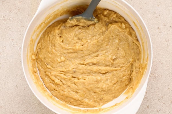 This Peanut Butter Banana Bread actually tastes like peanut butter! So soft, tender, and packed with both chocolate AND peanut butter chips. A white mixing bowl filled with thick, creamy banana bread batter being stirred with a gray spatula on a light countertop.