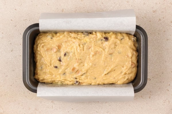 This Peanut Butter Banana Bread actually tastes like peanut butter! So soft, tender, and packed with both chocolate AND peanut butter chips. A loaf pan lined with parchment paper is filled with unbaked banana bread batter containing visible chocolate chips and peanut butter chips, sitting on a light-colored countertop.