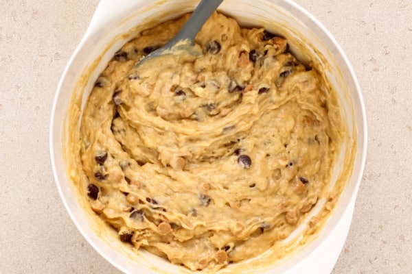 This Peanut Butter Banana Bread actually tastes like peanut butter! So soft, tender, and packed with both chocolate AND peanut butter chips. A white mixing bowl filled with thick banana bread batter containing chocolate chips and peanut butter chips, with a gray spatula resting inside.