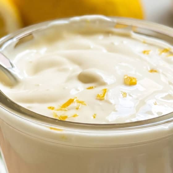 A glass of creamy garlic lemon aioli topped with lemon zest, with a small spoon inside. Slices of lemon are visible in the background on a striped cloth.