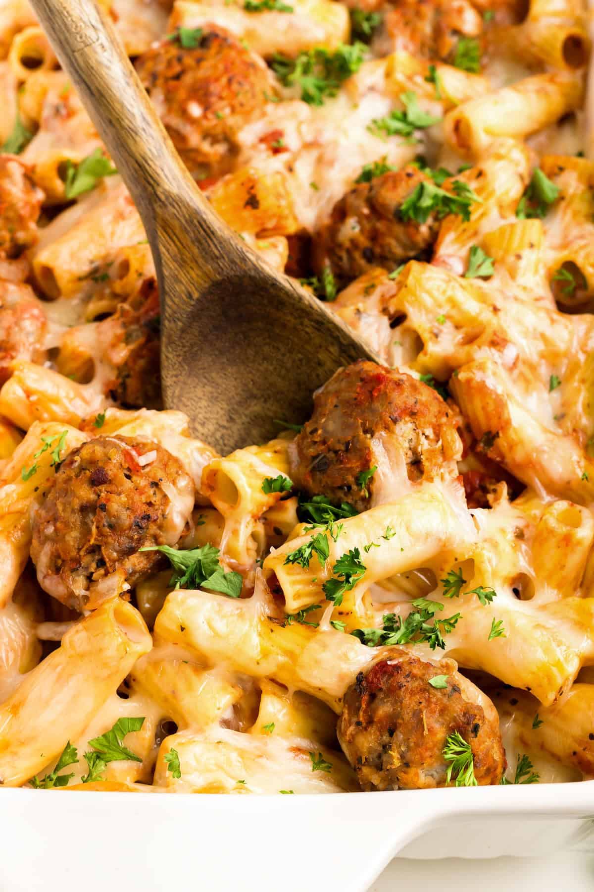 With seared meatballs, lots of cheese, and perfectly cooked saucy pasta (that's never mushy), this is the best Meatball Casserole recipe! A close-up of baked meatball casserole, covered in melted cheese and garnished with chopped parsley. A wooden spoon is scooping a serving from the dish.