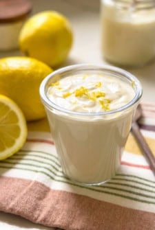 A glass filled with creamy lemon aioli topped with lemon zest, placed on a striped cloth next to whole and sliced lemons, with a jar in the background.
