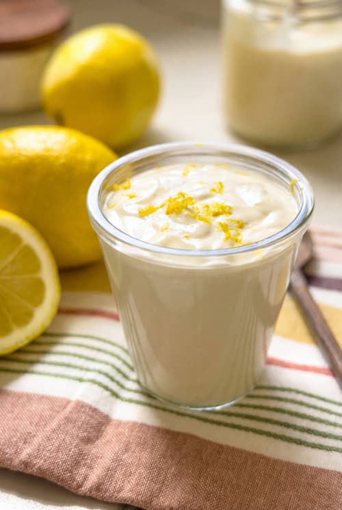 A glass filled with creamy lemon aioli topped with lemon zest, placed on a striped cloth next to whole and sliced lemons, with a jar in the background.