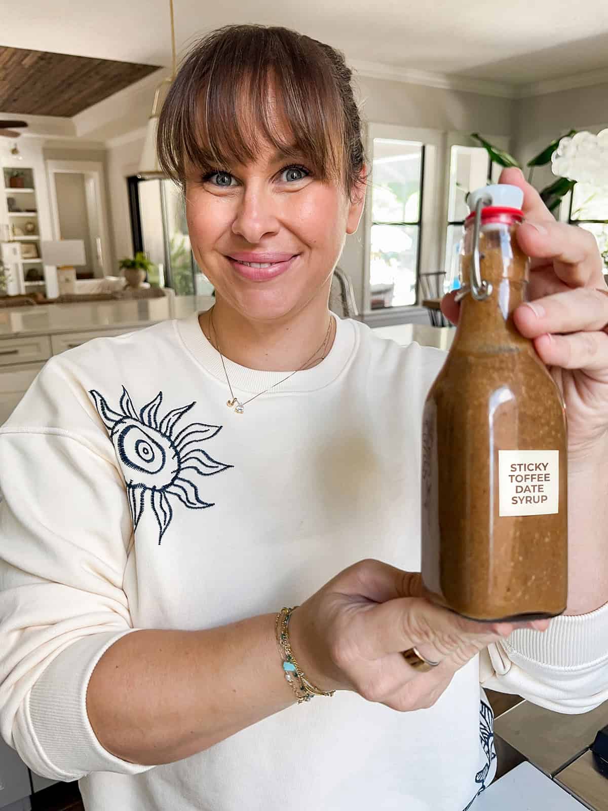 Sticky Toffee Date Coffee Syrup is made with 3 ingredients and NO cooking. Soak dates and toffee bits in hot espresso, blend, and enjoy! A smiling woman in a white sweater holds up a glass bottle of Sticky Toffee Date Coffee Syrup in a bright kitchen.