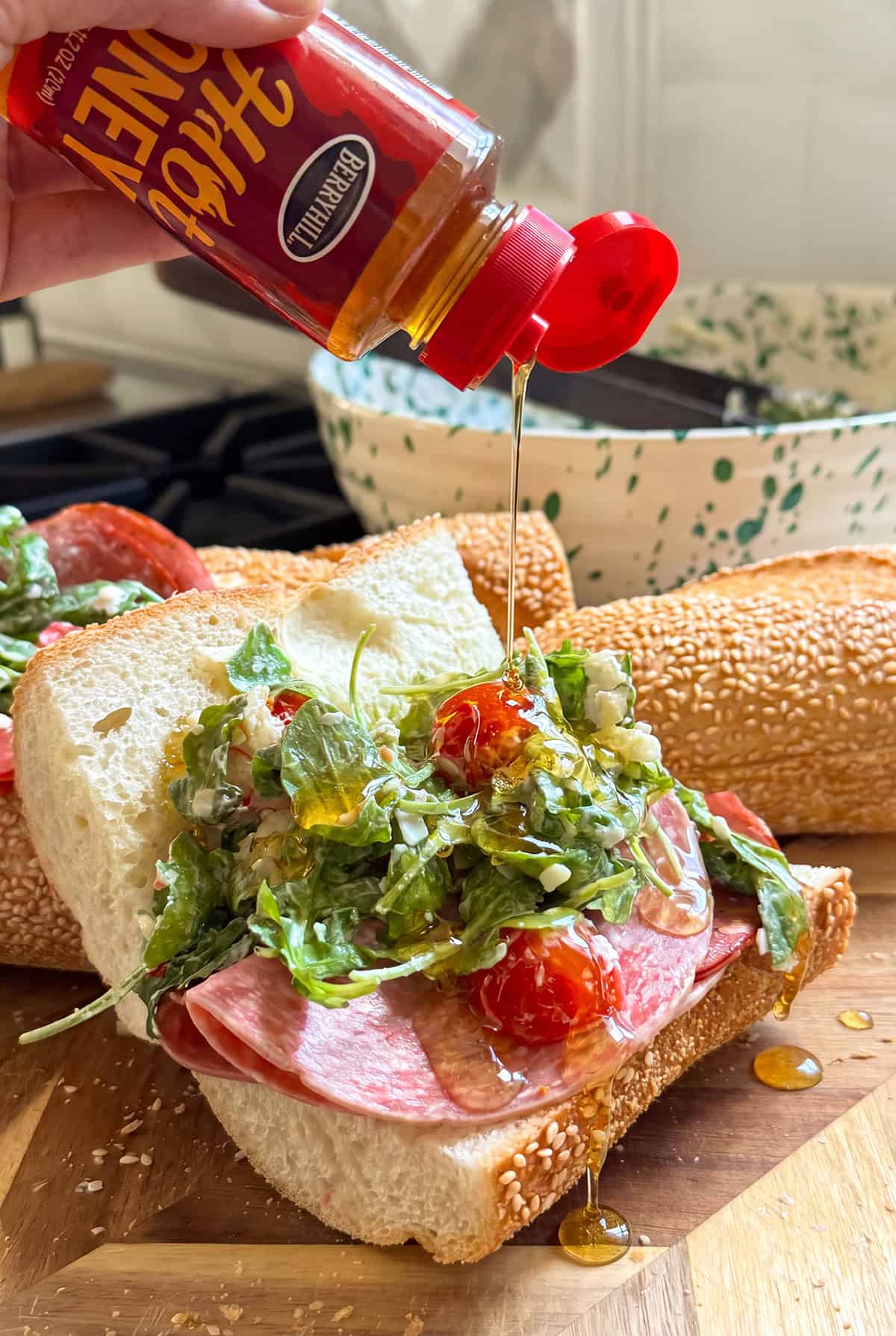 Is it a salad? Is it a sub? It's both. This Hot Honey Salami Sandwich is sweet, spicy, zesty, and salty in every bite. Ready in 10 minutes! A hand drizzles hot honey onto an open capicola and salami sandwich with arugula, tomatoes, and sesame seed bread. A loaf of bread and a bowl sit in the background on a wooden surface.