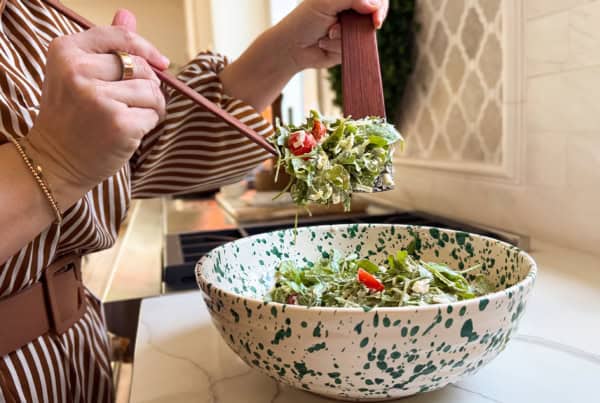 Is it a salad? Is it a sub? It's both. This Hot Honey Salami Sandwich is sweet, spicy, zesty, and salty in every bite. Ready in 10 minutes! A person tosses a fresh arugula salad with wooden utensils over a white bowl with green speckles on a kitchen counter.