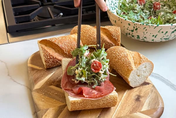 Is it a salad? Is it a sub? It's both. This Hot Honey Salami Sandwich is sweet, spicy, zesty, and salty in every bite. Ready in 10 minutes! A hand uses tongs to place a salad with arugula and cherry tomatoes on top of sliced sub bread with meat on a wooden cutting board.