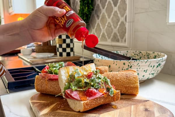 Is it a salad? Is it a sub? It's both. This Hot Honey Salami Sandwich is sweet, spicy, zesty, and salty in every bite. Ready in 10 minutes! A hand pours hot honey over two salami sandwiches filled with deli meats, white cheddar cheese, cherry tomatoes, and greens on a wooden cutting board.