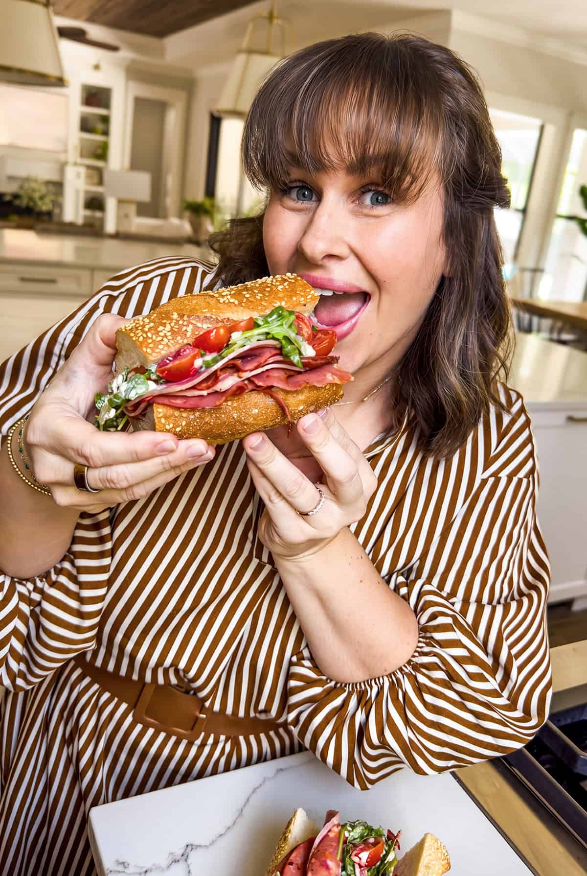 Is it a salad? Is it a sub? It's both. This Hot Honey Salami Sandwich is sweet, spicy, zesty, and salty in every bite. Ready in 10 minutes! A woman in a brown and white striped dress smiles and holds a large hot honey salami sandwich with deli meat, lettuce, and tomatoes in a bright kitchen. Another sandwich is on a plate in front of her.