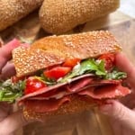 A person holds a hot honey salami sandwich filled with layers of deli meat, sliced cherry tomatoes, and fresh greens. Two pieces of sub bread are visible in the background on a wooden board.