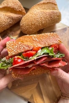 A person holds a hot honey salami sandwich filled with layers of deli meat, sliced cherry tomatoes, and fresh greens. Two pieces of sub bread are visible in the background on a wooden board.