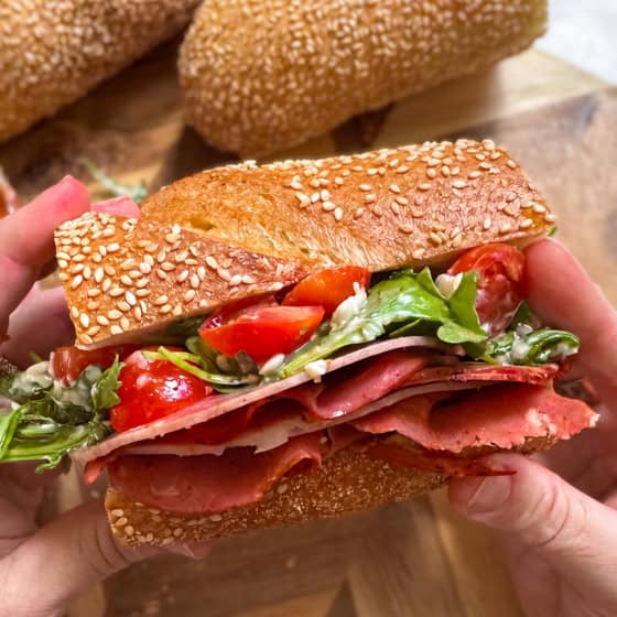 A person holds a hot honey salami sandwich filled with layers of deli meat, sliced cherry tomatoes, and fresh greens. Two pieces of sub bread are visible in the background on a wooden board.