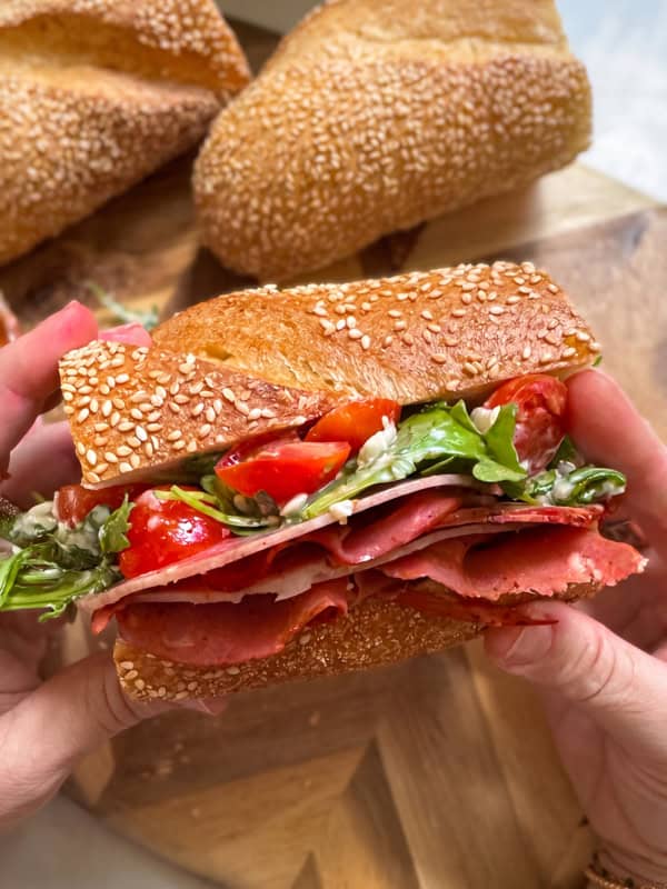 A person holds a hot honey salami sandwich filled with layers of deli meat, sliced cherry tomatoes, and fresh greens. Two pieces of sub bread are visible in the background on a wooden board.
