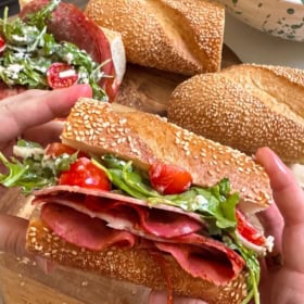 A hand holds a Hot Honey Italian Sub roll filled with salami, arugula, sliced tomatoes, and creamy dressing. Two more salami sandwiches and a bowl of salad are on a wooden surface.