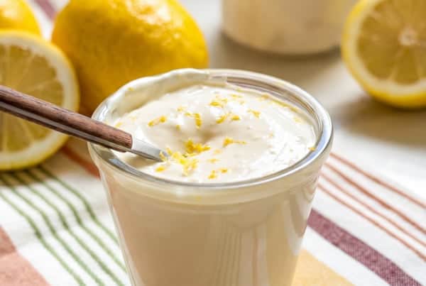 Creamy lemon aioli recipe with a bright, tangy twist! The secret is sour cream. Whisk it up in 5 minutes and keep it in the fridge for 2 weeks. A glass of creamy garlic lemon aioli topped with lemon zest with a small spoon inside. Fresh lemons and lemon halves are in the background on a striped cloth.