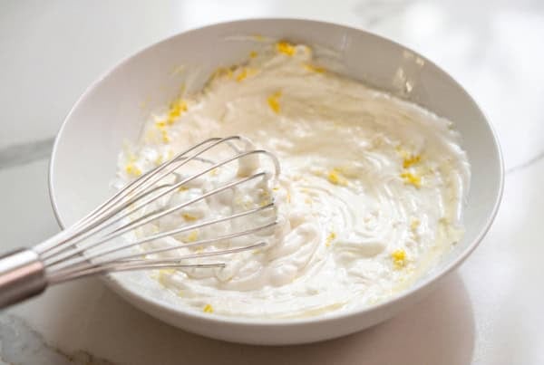 Creamy lemon aioli recipe with a bright, tangy twist! The secret is sour cream. Whisk it up in 5 minutes and keep it in the fridge for 2 weeks. A metal whisk rests in a white bowl containing a creamy mixture with visible yellow lemon zest on a white countertop.