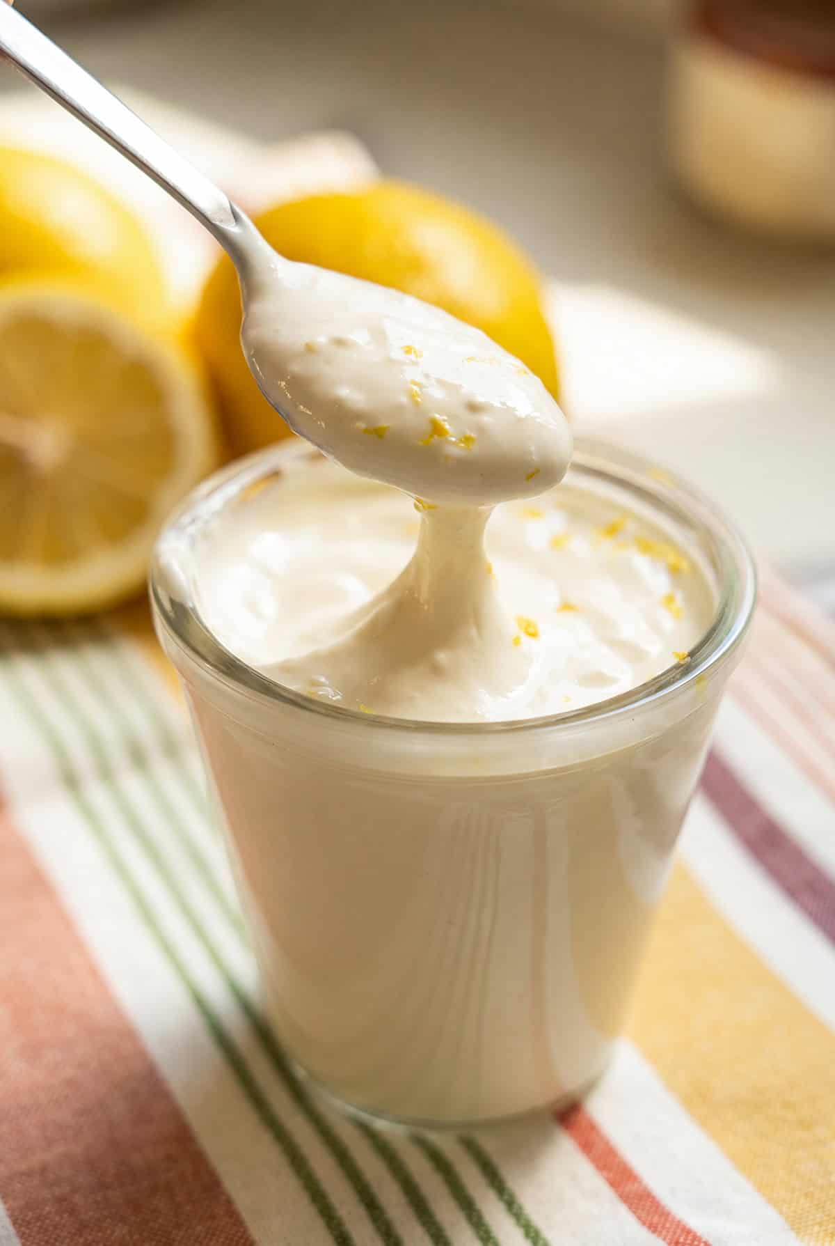 Creamy lemon aioli recipe with a bright, tangy twist! The secret is sour cream. Whisk it up in 5 minutes and keep it in the fridge for 2 weeks. A spoon lifts a spoonful of garlicky lemon aioli with lemon zest from a glass jar. Sliced lemons and whole lemons are in the background on a colorful striped cloth.
