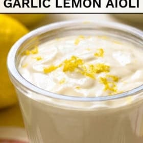 A glass container filled with creamy garlic lemon aioli, topped with lemon zest. Whole lemons are visible in the background. The text above reads Easy 5-Ingredient Garlic Lemon Aioli and below, The Novice Chef.