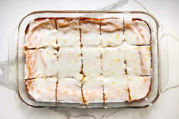 These lemon brownies are thick, chewy, packed with real lemon flavor, and topped with a sweet-tart glaze. Ready for the oven in 10 minutes! A glass baking dish filled with rectangular lemon blondies, topped with a glossy white glaze and sprinkled with lemon zest, cut into 15 squares on a white marble surface.