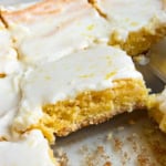 A close-up of lemon brownies cut into squares in a glass baking dish, with a thin layer of white lemon glaze on top. One piece is slightly lifted from the dish.