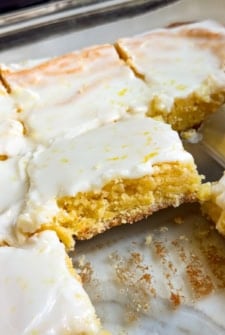 A close-up of lemon brownies cut into squares in a glass baking dish, with a thin layer of white lemon glaze on top. One piece is slightly lifted from the dish.