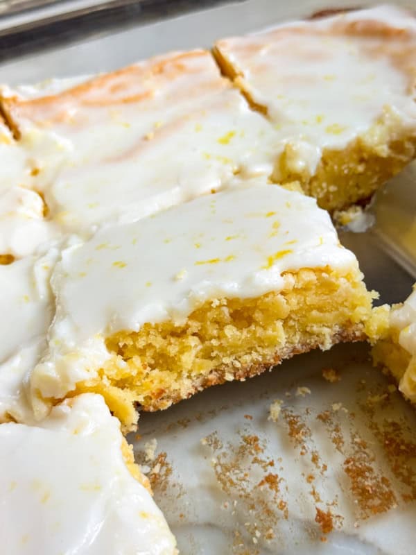 A close-up of lemon brownies cut into squares in a glass baking dish, with a thin layer of white lemon glaze on top. One piece is slightly lifted from the dish.