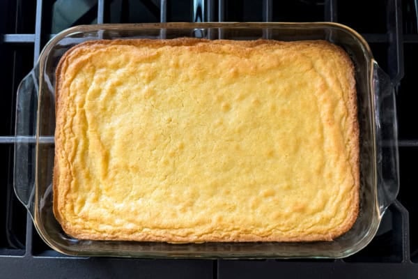 These lemon brownies are thick, chewy, packed with real lemon flavor, and topped with a sweet-tart glaze. Ready for the oven in 10 minutes! A 9x13 glass pan filled with freshly baked lemon brownies is resting on a black stovetop. They have a lightly cracked surface and slightly raised, browned edges.