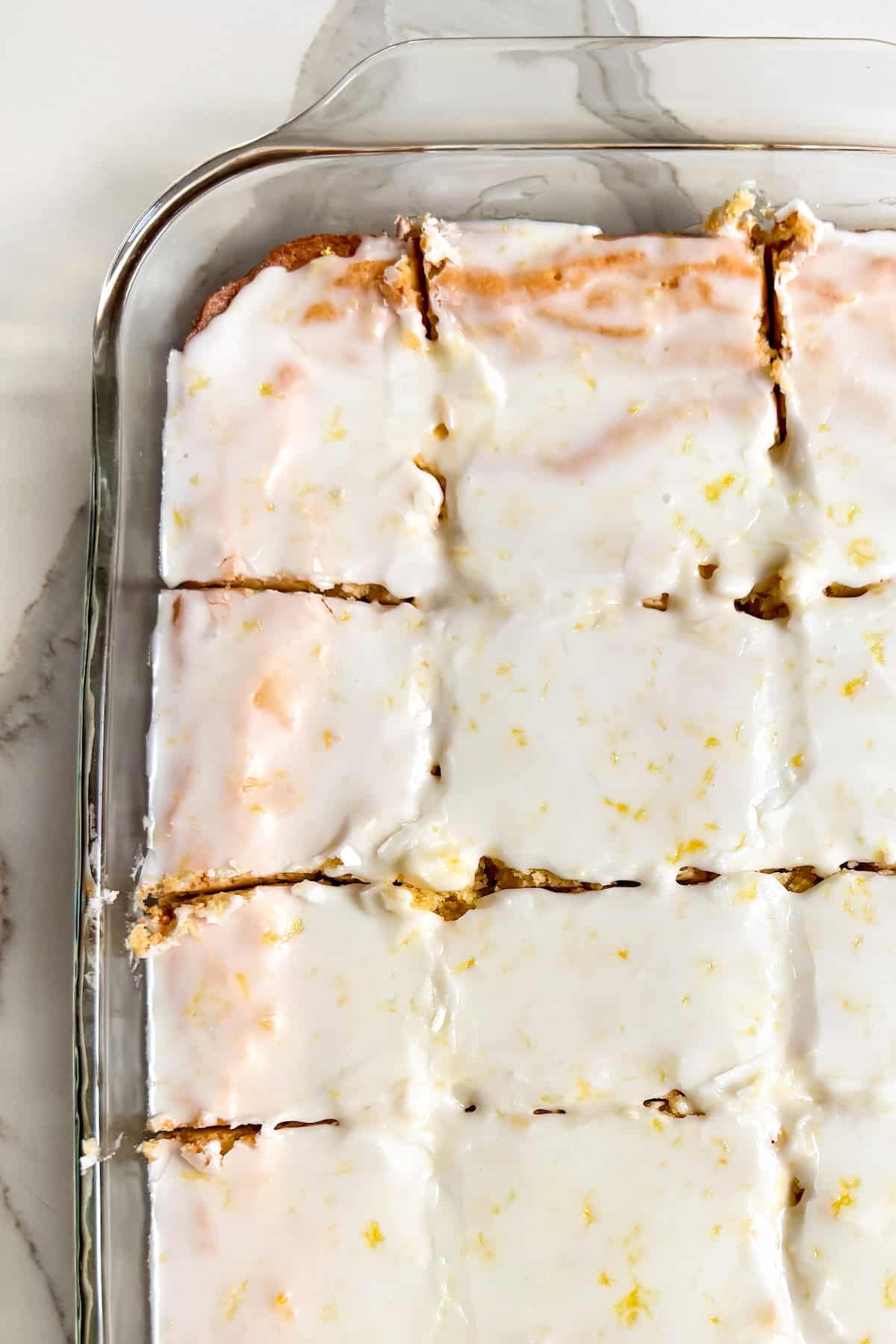 These lemon brownies are thick, chewy, packed with real lemon flavor, and topped with a sweet-tart glaze. Ready for the oven in 10 minutes! A glass baking dish filled with rectangular lemon brownies covered in a glossy white lemon glaze, cut into squares and placed on a marble countertop.