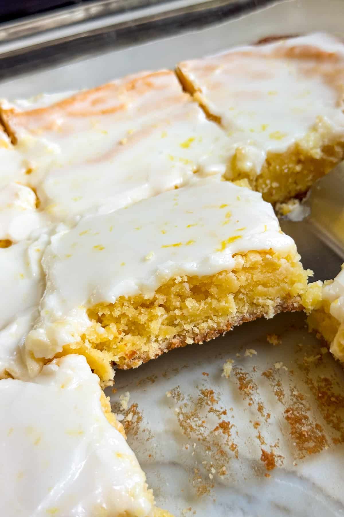 These lemon brownies are thick, chewy, packed with real lemon flavor, and topped with a sweet-tart glaze. Ready for the oven in 10 minutes! A close-up of lemon brownies cut into squares in a glass baking dish, with a thin layer of white lemon glaze on top. One piece is slightly lifted from the dish.